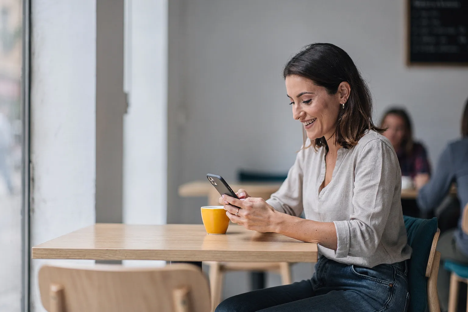 Mujer sonriendo mientras escribe una reseña en el móvil sentada en una cafetería luminosa